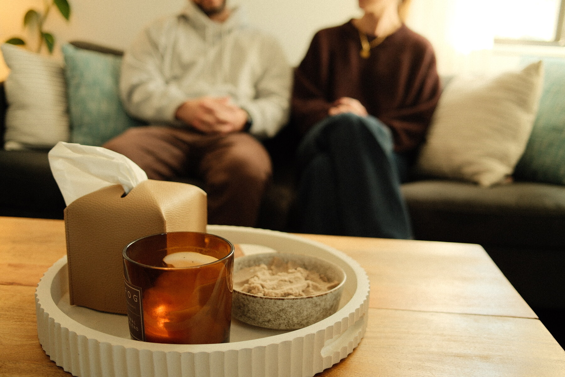A close detail of a candle and tissue box on a coffee table, with two clients seated together on the couch in soft focus.