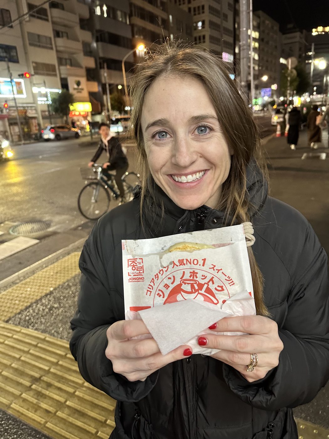 Jess Mansell smiling on a city street at night in Japan, holding a local street food snack.